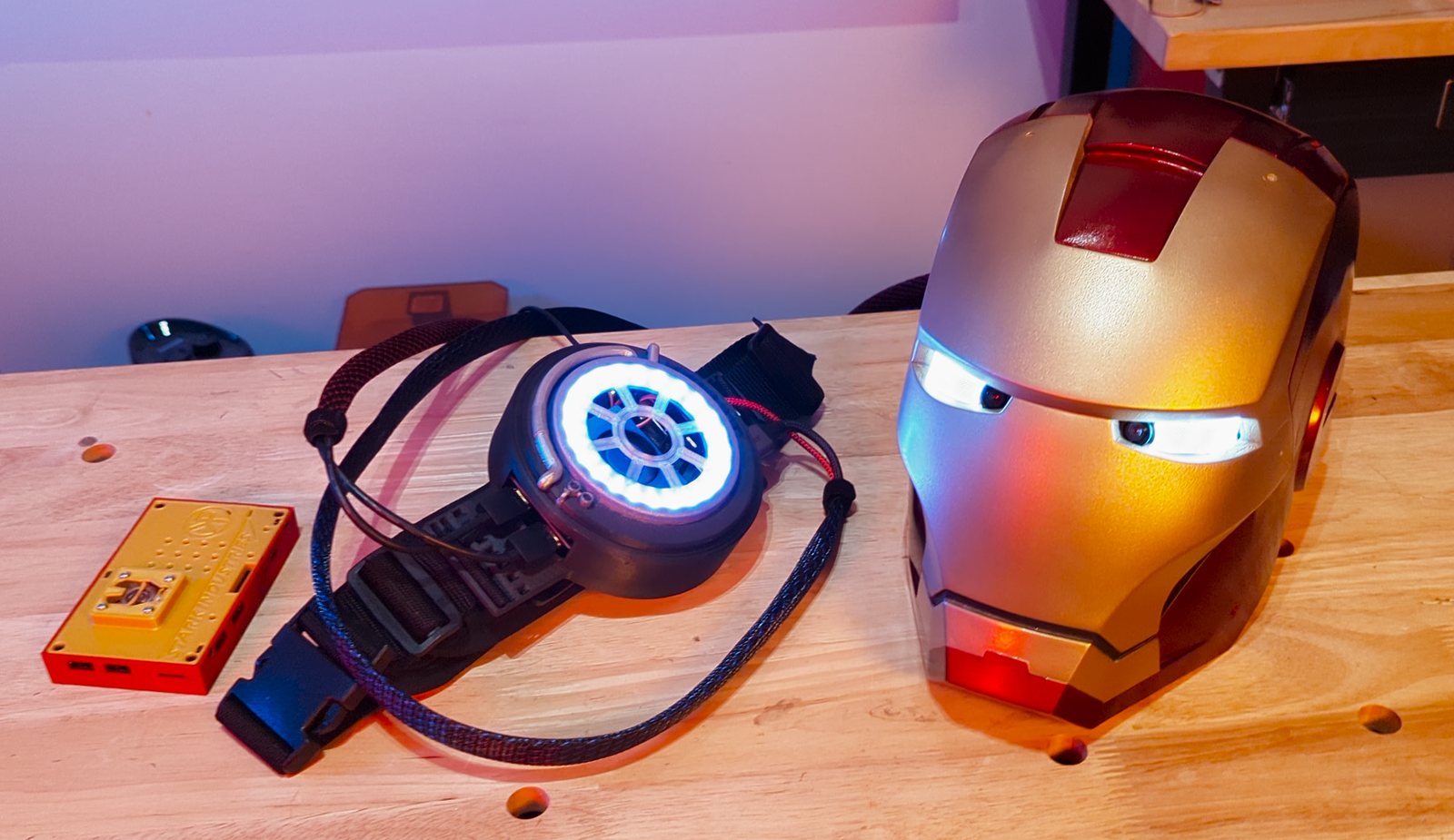 A finished red-and-gold Iron Man helmet with its eyes lit, on the bench beside a glowing arc reactor chest piece and a Stark Industries control box